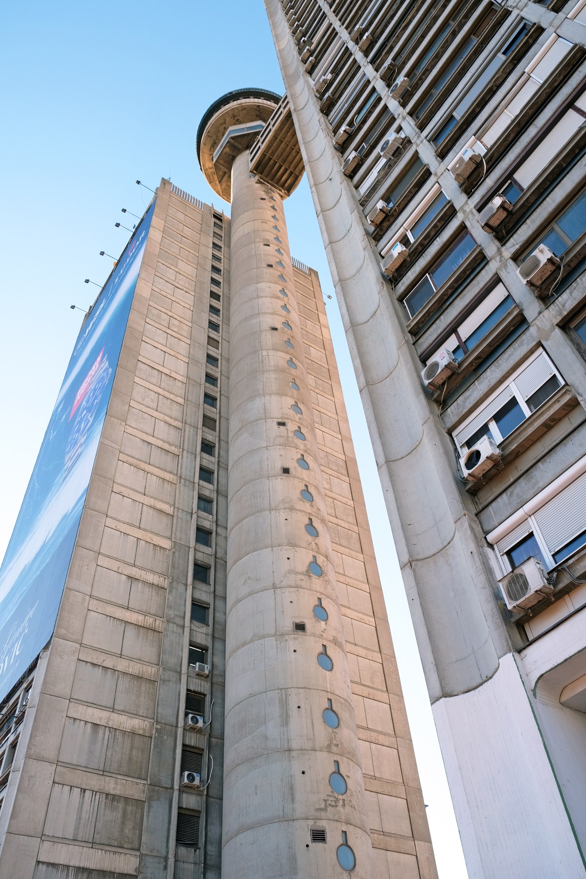 Low-angle view of Genex Tower’s concrete cylindrical core and repeated apartment balconies against a blue sky.