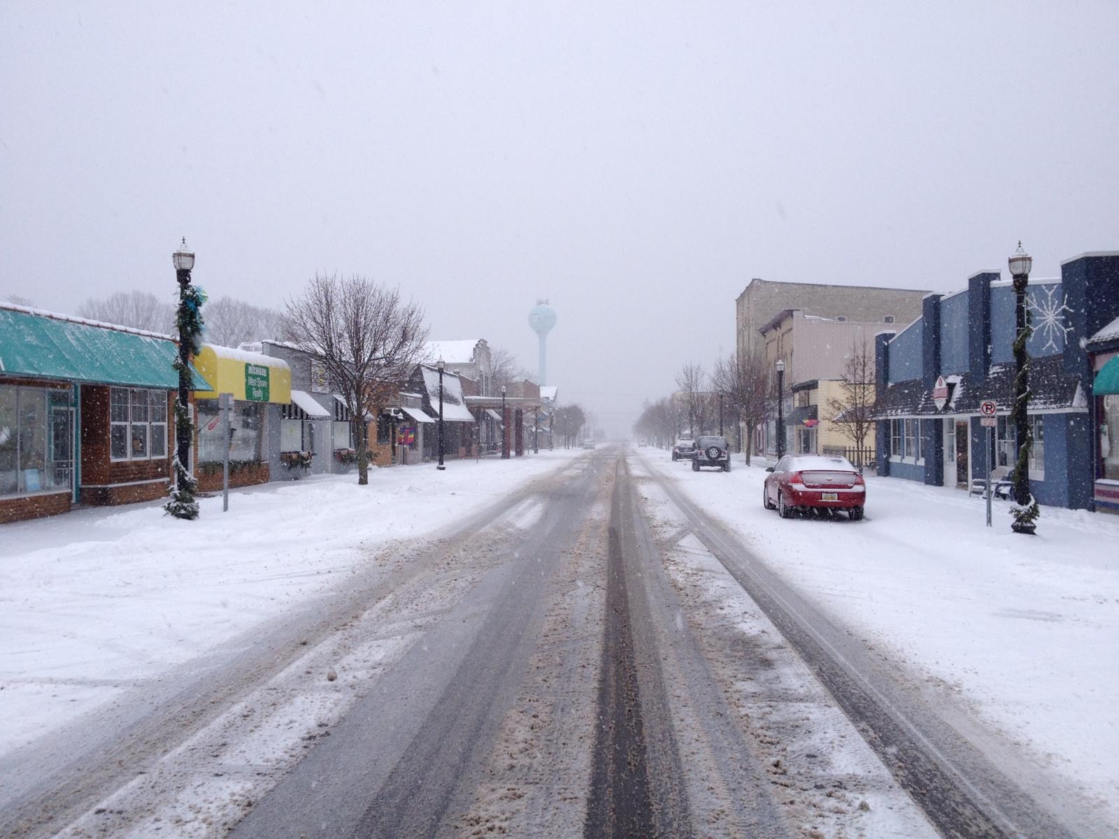 Snow-covered main street in Pentwater, Michigan, with modest storefronts and a red parked car.