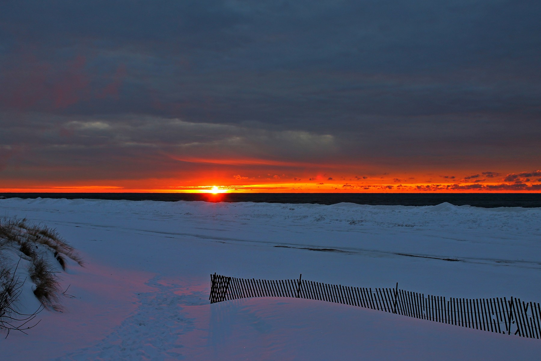 Winter sunset over Lake Michigan, with snow-covered dunes and a weathered wooden sand fence.